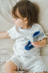 Baby girl in Avauma Mellow Days Vanilla Herb set, sitting on bed and holding a blue toy. Short sleeve top and shorts with dainty green flower prints. Baby girl in Avauma Mellow Days Vanilla Herb set, sitting on bed and holding a blue toy. Short sleeve top and shorts with dainty green flower prints.