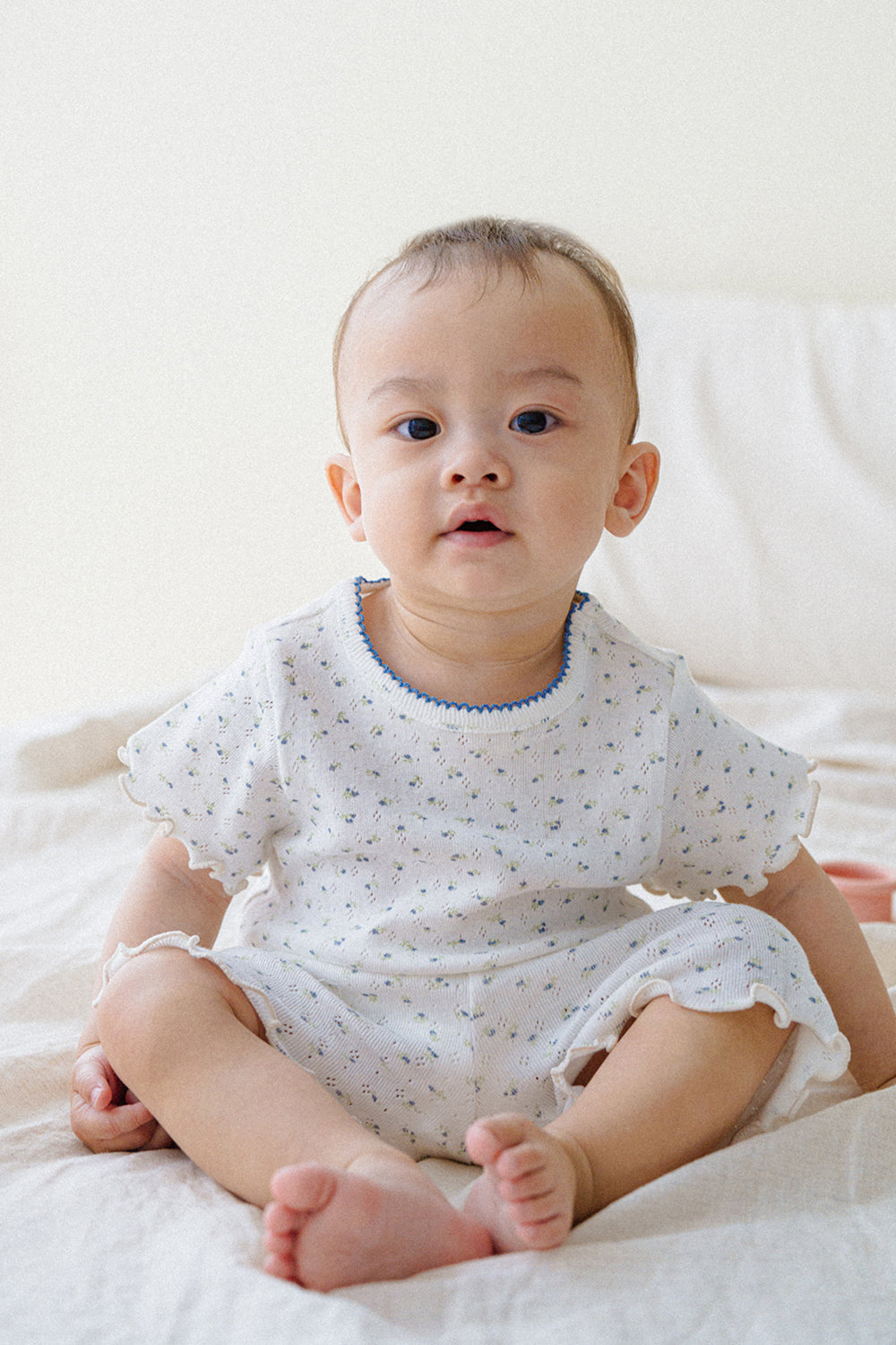 Baby boy in Mellow Days Vanilla Herb baby set by Avauma, sitting on a bed. Adorable summer pajamas in white with tiny green floral design. Baby boy in Mellow Days Vanilla Herb baby set by Avauma, sitting on a bed. Adorable summer pajamas in white with tiny green floral design.