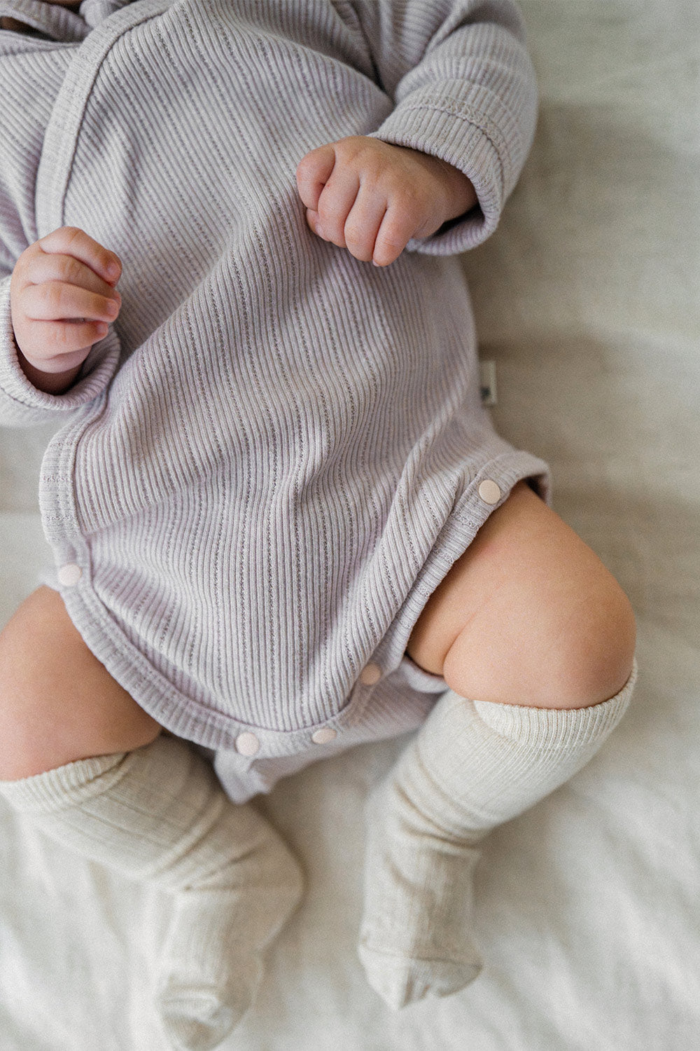 Detailed view of the Avauma Selina Babysuit in lavender haze, focusing on the wrap design, snap closures, and stretchy ribbed cotton that supports easy movement for active babies. Detailed view of the Avauma Selina Babysuit in lavender haze, focusing on the wrap design, snap closures, and stretchy ribbed cotton that supports easy movement for active babies.