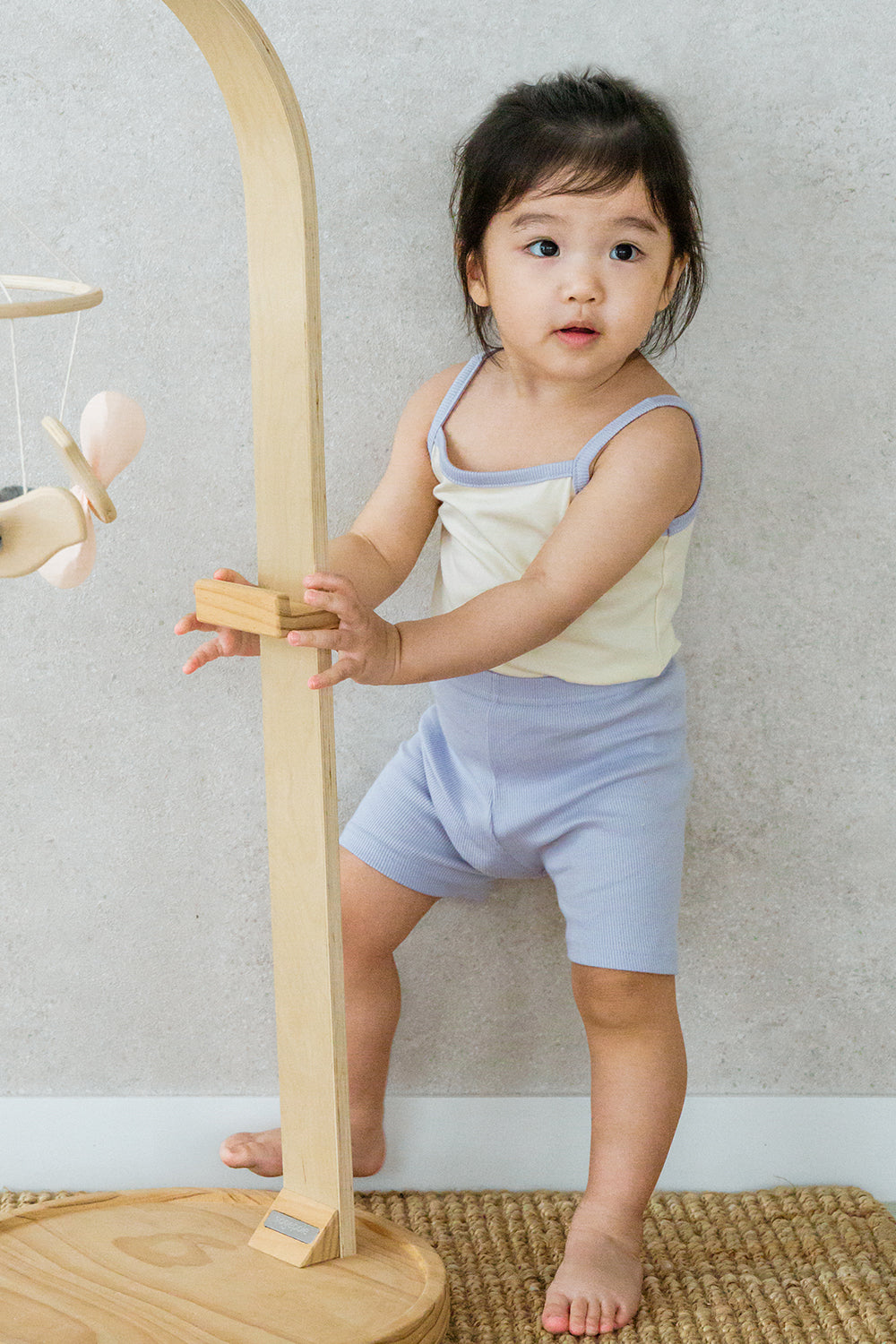 Toddler girl in AVAUMA Laila Day lilac cream sleeveless set, standing and holding wooden toy Toddler girl in AVAUMA Laila Day lilac cream sleeveless set, standing and holding wooden toy