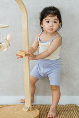 Toddler girl in AVAUMA Laila Day lilac cream sleeveless set, standing and holding wooden toy Toddler girl in AVAUMA Laila Day lilac cream sleeveless set, standing and holding wooden toy