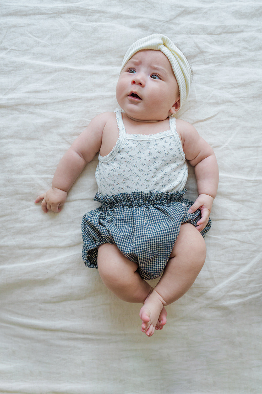 Baby wearing Avauma Lily Sleeveless Bodysuit Cream Blue. Infant in a white floral print bodysuit, dark blue checkered bloomers, and a white headband.
Baby wearing Avauma Lily Sleeveless Bodysuit Cream Blue. Infant in a white floral print bodysuit, dark blue checkered bloomers, and a white headband.