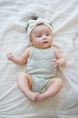 Adorable baby model in Avauma Lily Sleeveless Bodysuit Sage Flower. Infant lying down in a soft sage green floral print bodysuit with a twisted white headband. Adorable baby model in Avauma Lily Sleeveless Bodysuit Sage Flower. Infant lying down in a soft sage green floral print bodysuit with a twisted white headband.