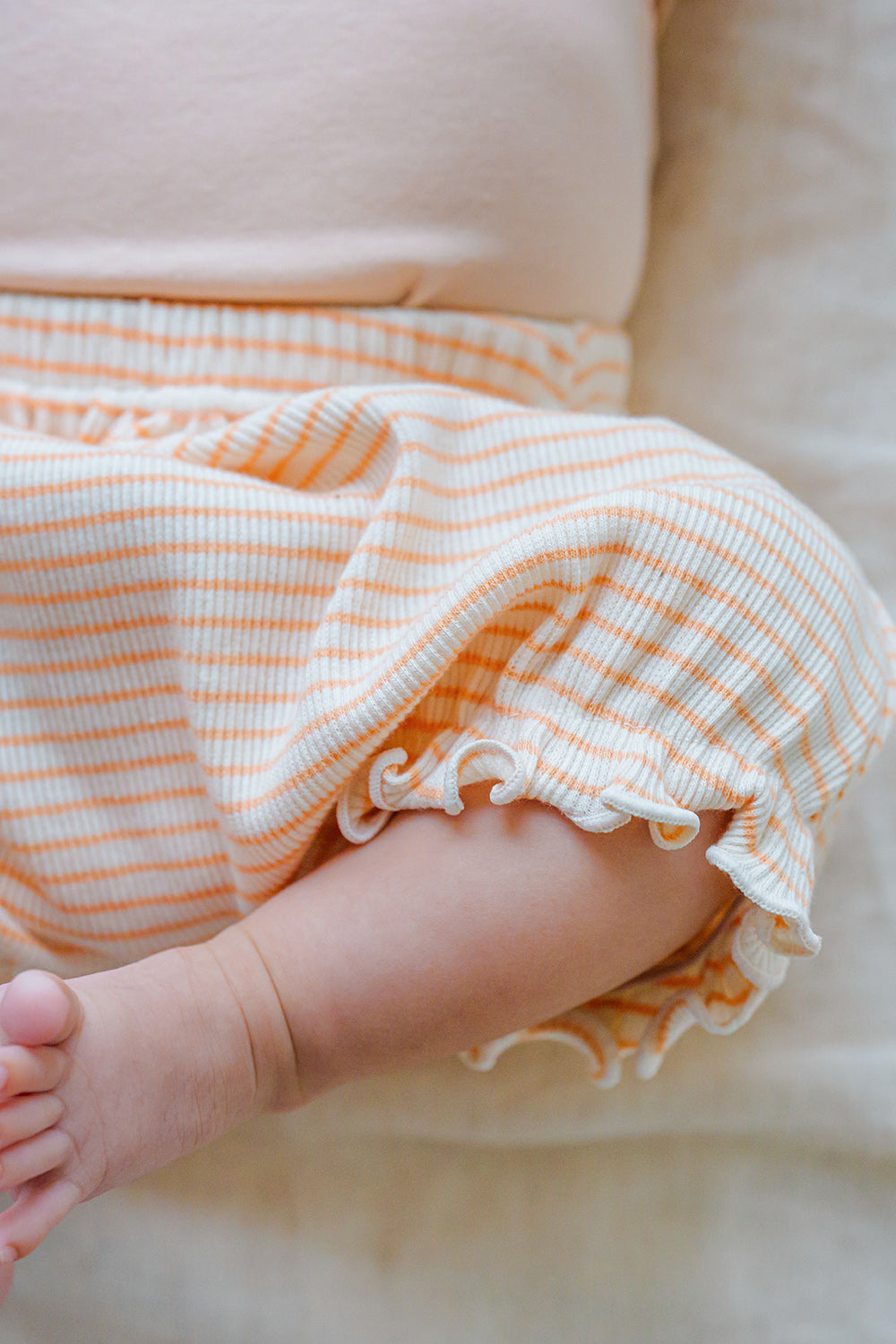 Close-up of Avauma Mellow Stripe Set Peach Macaron. Detail of baby's arm and leg in the striped bloomers with ruffled trim, showing fabric texture. Close-up of Avauma Mellow Stripe Set Peach Macaron. Detail of baby's arm and leg in the striped bloomers with ruffled trim, showing fabric texture.