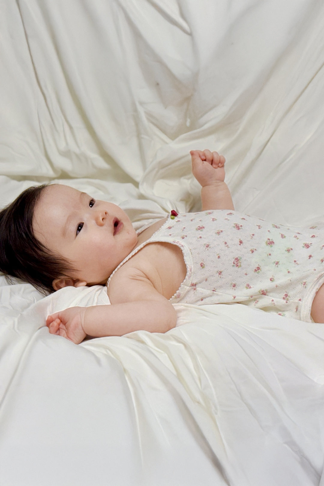 Baby wearing Avauma Summer Picot Sleeveless Bodysuit Rosebell. Infant lying on back, looking up, in a comfortable white floral print romper. Baby wearing Avauma Summer Picot Sleeveless Bodysuit Rosebell. Infant lying on back, looking up, in a comfortable white floral print romper.
