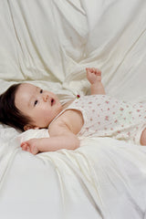 Baby wearing Avauma Summer Picot Sleeveless Bodysuit Rosebell. Infant lying on back, looking up, in a comfortable white floral print romper. Baby wearing Avauma Summer Picot Sleeveless Bodysuit Rosebell. Infant lying on back, looking up, in a comfortable white floral print romper.