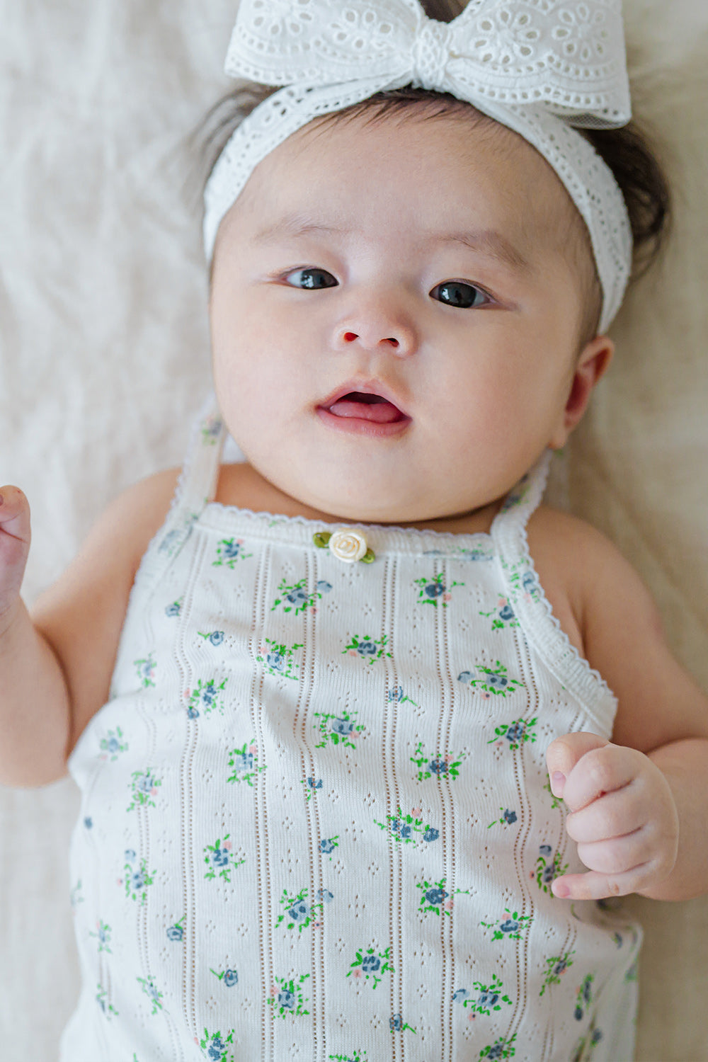 Cute baby face in Avauma Summer Picot Sleeveless Bodysuit Blue Garden Mint. Infant looking at camera, wearing floral romper and white lace bow headband. Cute baby face in Avauma Summer Picot Sleeveless Bodysuit Blue Garden Mint. Infant looking at camera, wearing floral romper and white lace bow headband.