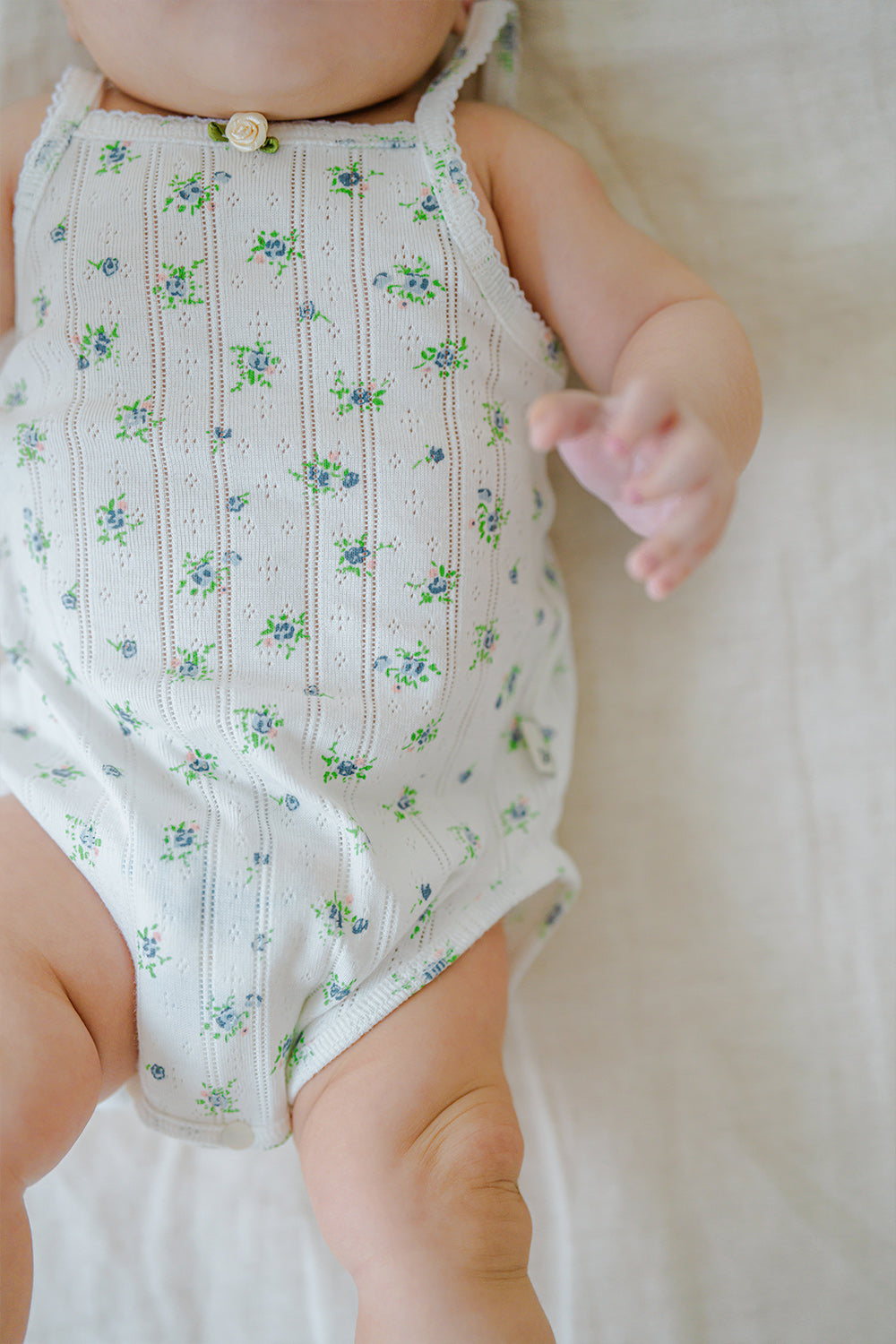 Close-up of Avauma Summer Picot Sleeveless Bodysuit Blue Garden Mint. Detail of white romper with blue and green floral pattern on baby's torso. Close-up of Avauma Summer Picot Sleeveless Bodysuit Blue Garden Mint. Detail of white romper with blue and green floral pattern on baby's torso.