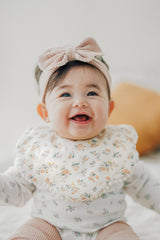 Smiling baby wearing Avauma Mellow Ribbon Band in Baby Blush and floral top
 Smiling baby wearing Avauma Mellow Ribbon Band in Baby Blush and floral top