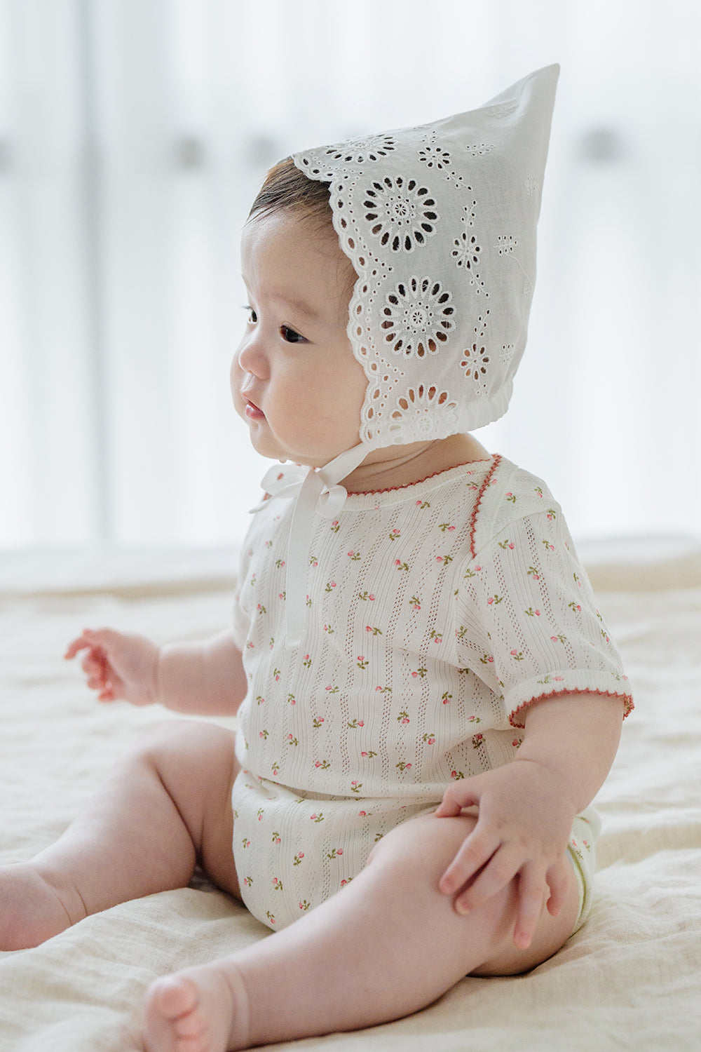 Baby girl wearing Avauma Clare Bonnet in Cotton Cream with circular embroidered eyelets, sitting on a soft beige bed Baby girl wearing Avauma Clare Bonnet in Cotton Cream with circular embroidered eyelets, sitting on a soft beige bed