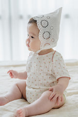 Baby girl wearing Avauma Clare Bonnet in Cotton Cream with circular embroidered eyelets, sitting on a soft beige bed Baby girl wearing Avauma Clare Bonnet in Cotton Cream with circular embroidered eyelets, sitting on a soft beige bed