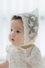 Baby girl wearing Avauma Clare Bonnet in Ivory Race, showing delicate floral lace and satin ribbon detail while held by caregiver Baby girl wearing Avauma Clare Bonnet in Ivory Race, showing delicate floral lace and satin ribbon detail while held by caregiver