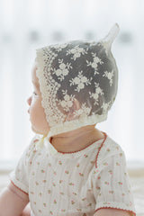 Baby in Avauma Clare Bonnet - Ivory Race with vintage-style floral lace and soft ivory outfit, looking to the side Baby in Avauma Clare Bonnet - Ivory Race with vintage-style floral lace and soft ivory outfit, looking to the side