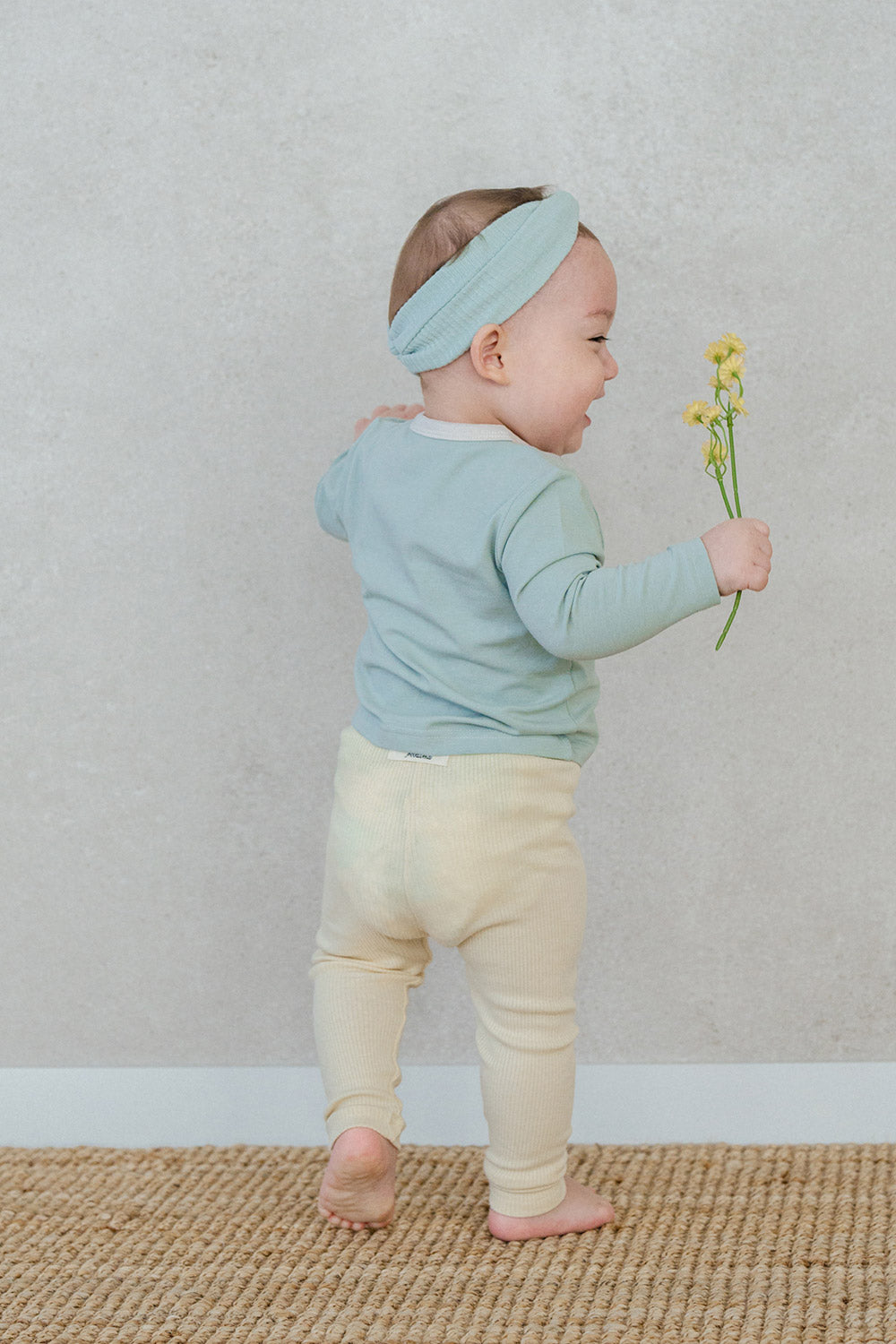Back view of a smiling baby holding yellow flowers – highlighting the comfortable waistband of the Avauma Cloud Ribbed Setup in Sage Leaf for unrestricted movement Back view of a smiling baby holding yellow flowers – highlighting the comfortable waistband of the Avauma Cloud Ribbed Setup in Sage Leaf for unrestricted movement