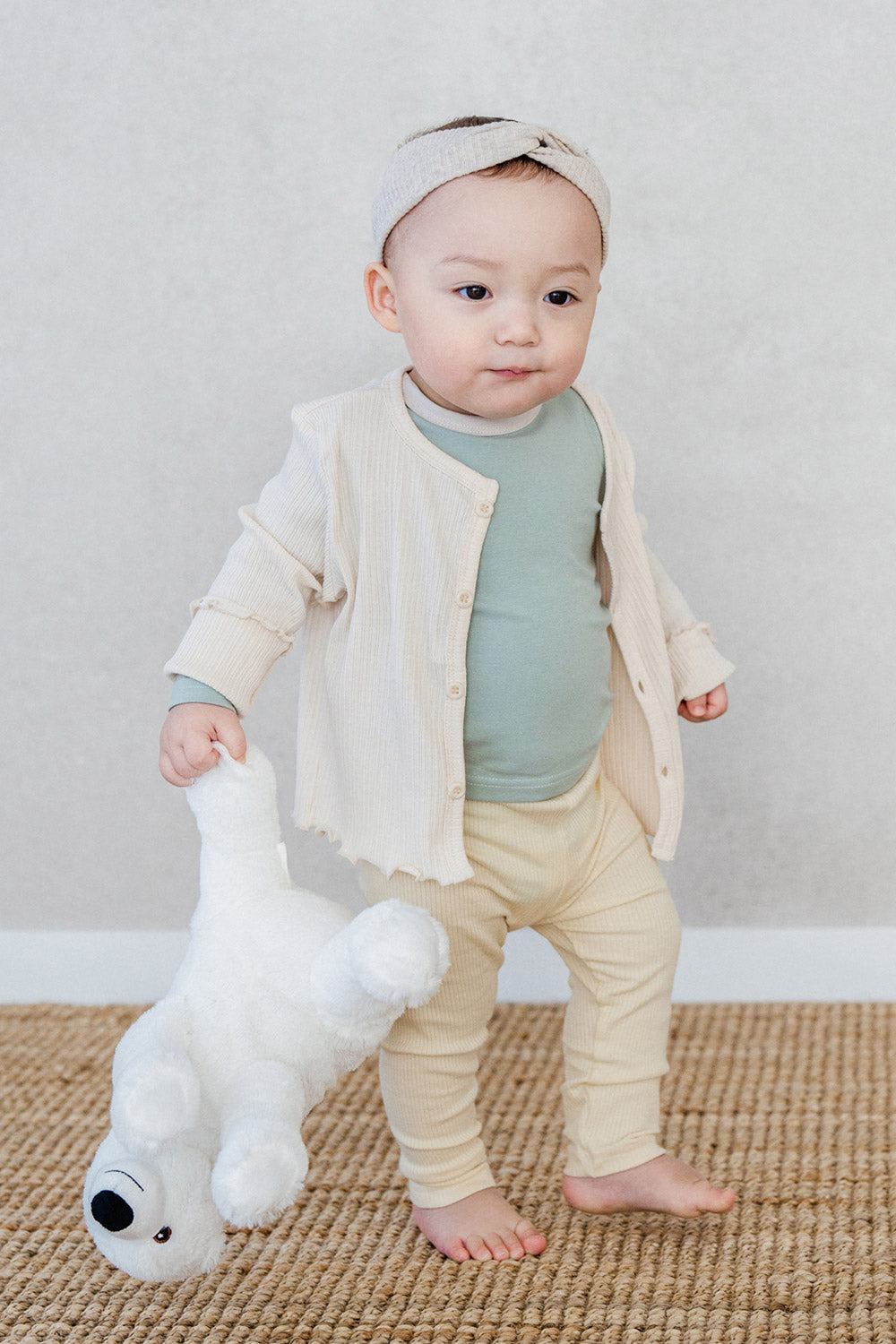 Baby standing with a stuffed toy – wearing a cream-colored cardigan over the Avauma Cloud Ribbed Setup in Sage Leaf for a warm, cozy look Baby standing with a stuffed toy – wearing a cream-colored cardigan over the Avauma Cloud Ribbed Setup in Sage Leaf for a warm, cozy look