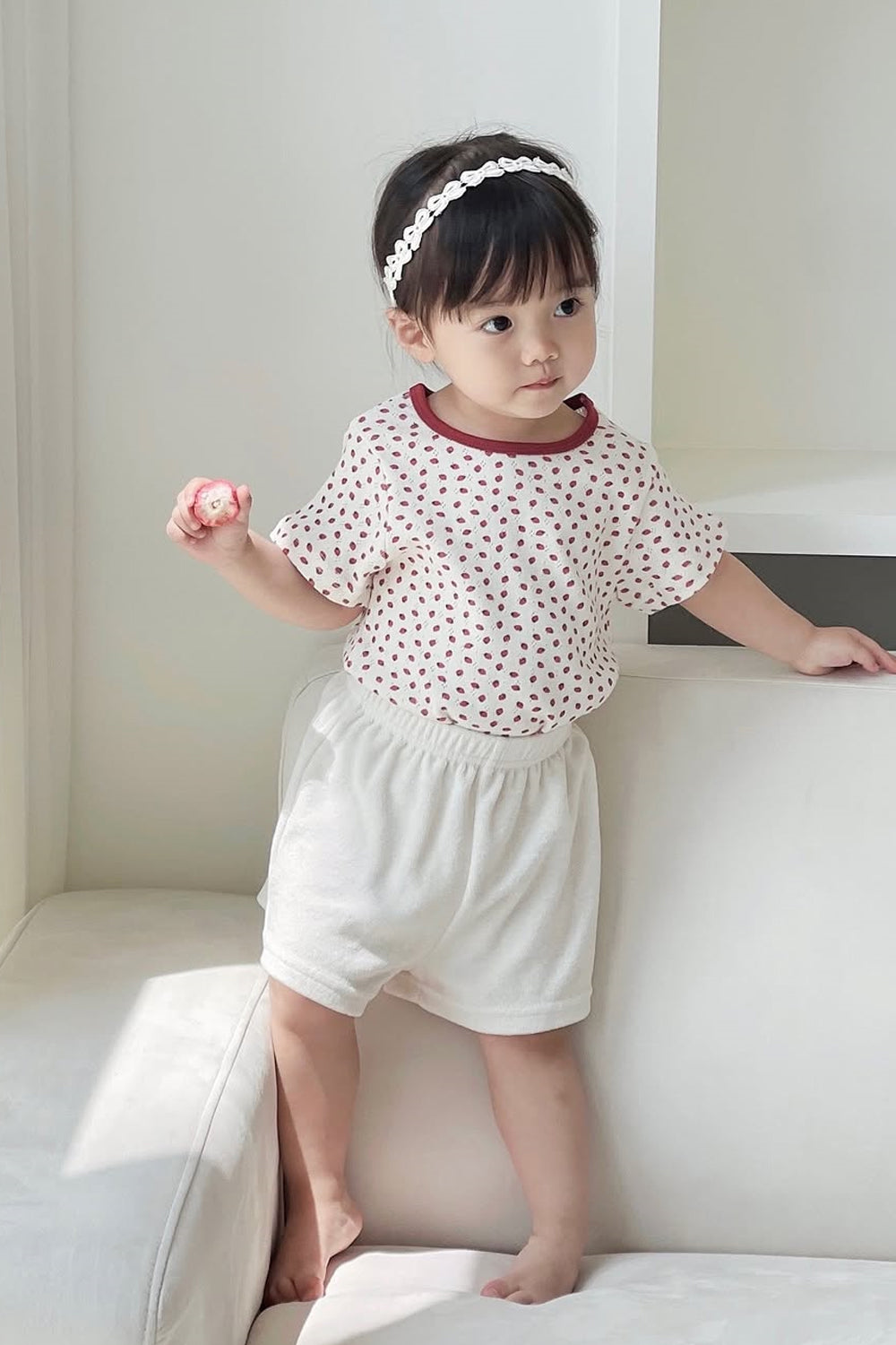 Adorable toddler girl standing on sofa in Avauma Mellow Pop Ivory Berry clothing, bright natural setting Adorable toddler girl standing on sofa in Avauma Mellow Pop Ivory Berry clothing, bright natural setting