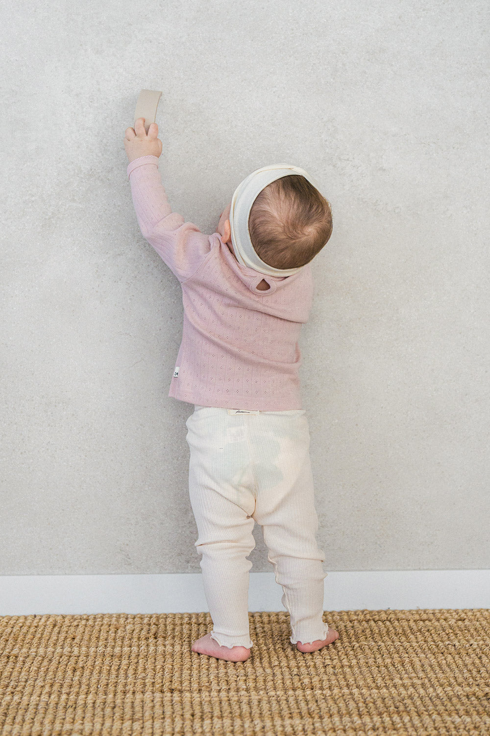 Back view of a baby standing against the wall – highlighting the back button detail and ruffle accents on the ribbed leggings in the Avauma Peach Glow Set in Pale Peach Back view of a baby standing against the wall – highlighting the back button detail and ruffle accents on the ribbed leggings in the Avauma Peach Glow Set in Pale Peach