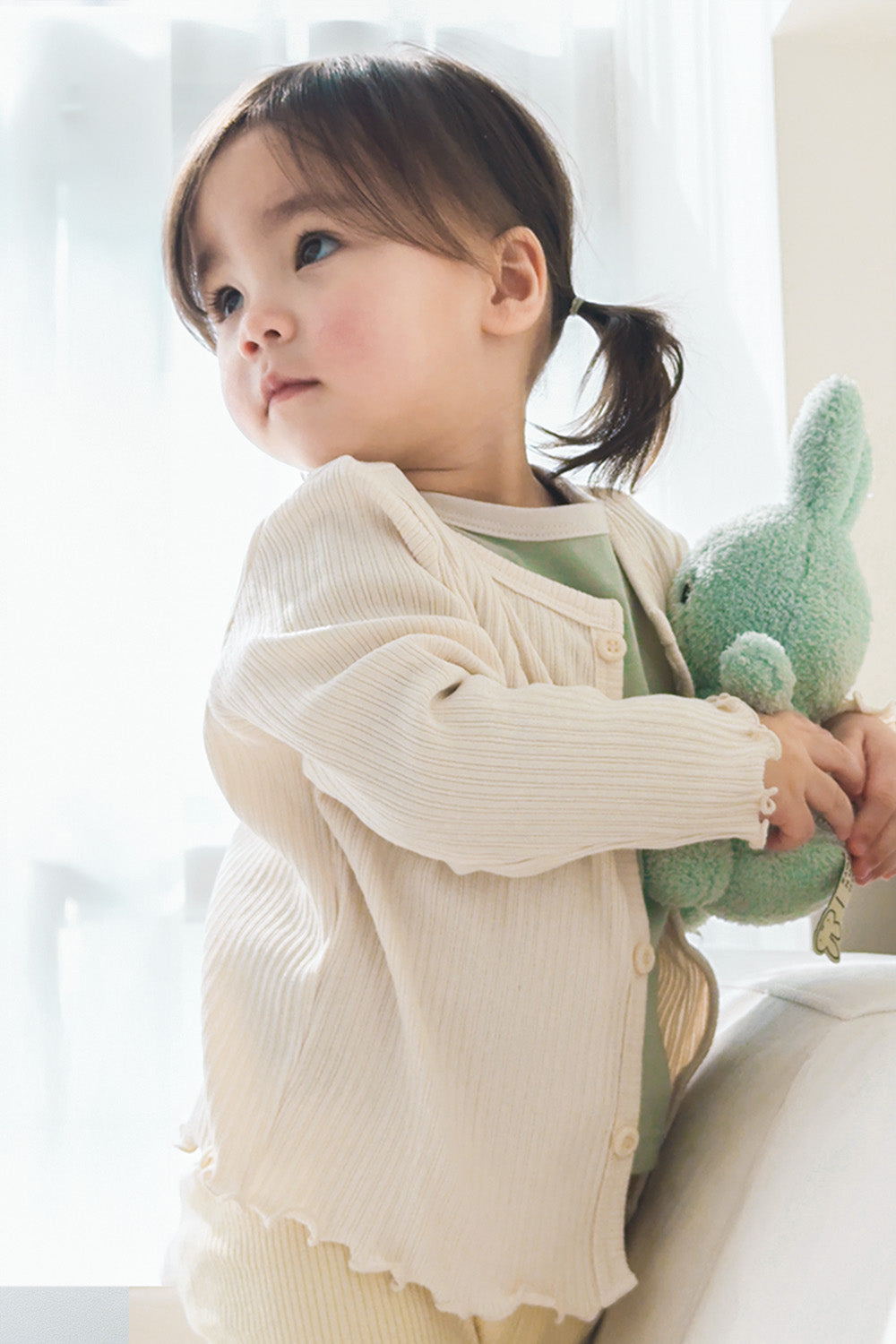 Toddler girl looking up in Avauma ribbed cardigan, soft natural off white cotton fabric Toddler girl looking up in Avauma ribbed cardigan, soft natural off white cotton fabric