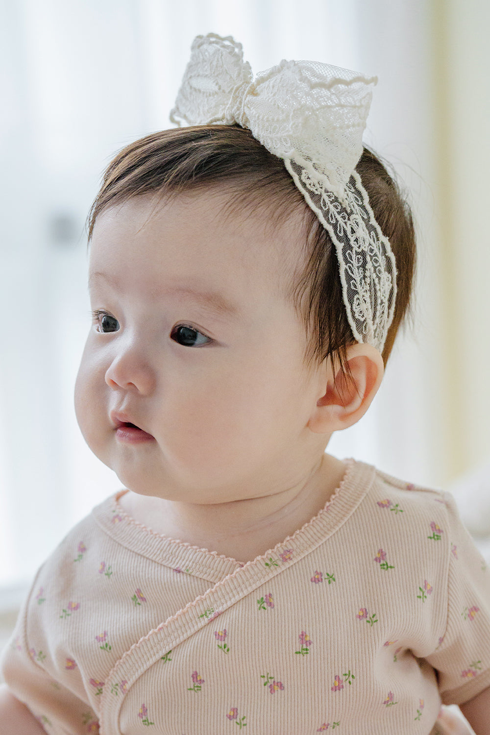 Baby girl wearing Avauma Rumiere Ribbon Band and pink floral wrap bodysuit, looking to the side indoors Baby girl wearing Avauma Rumiere Ribbon Band and pink floral wrap bodysuit, looking to the side indoors