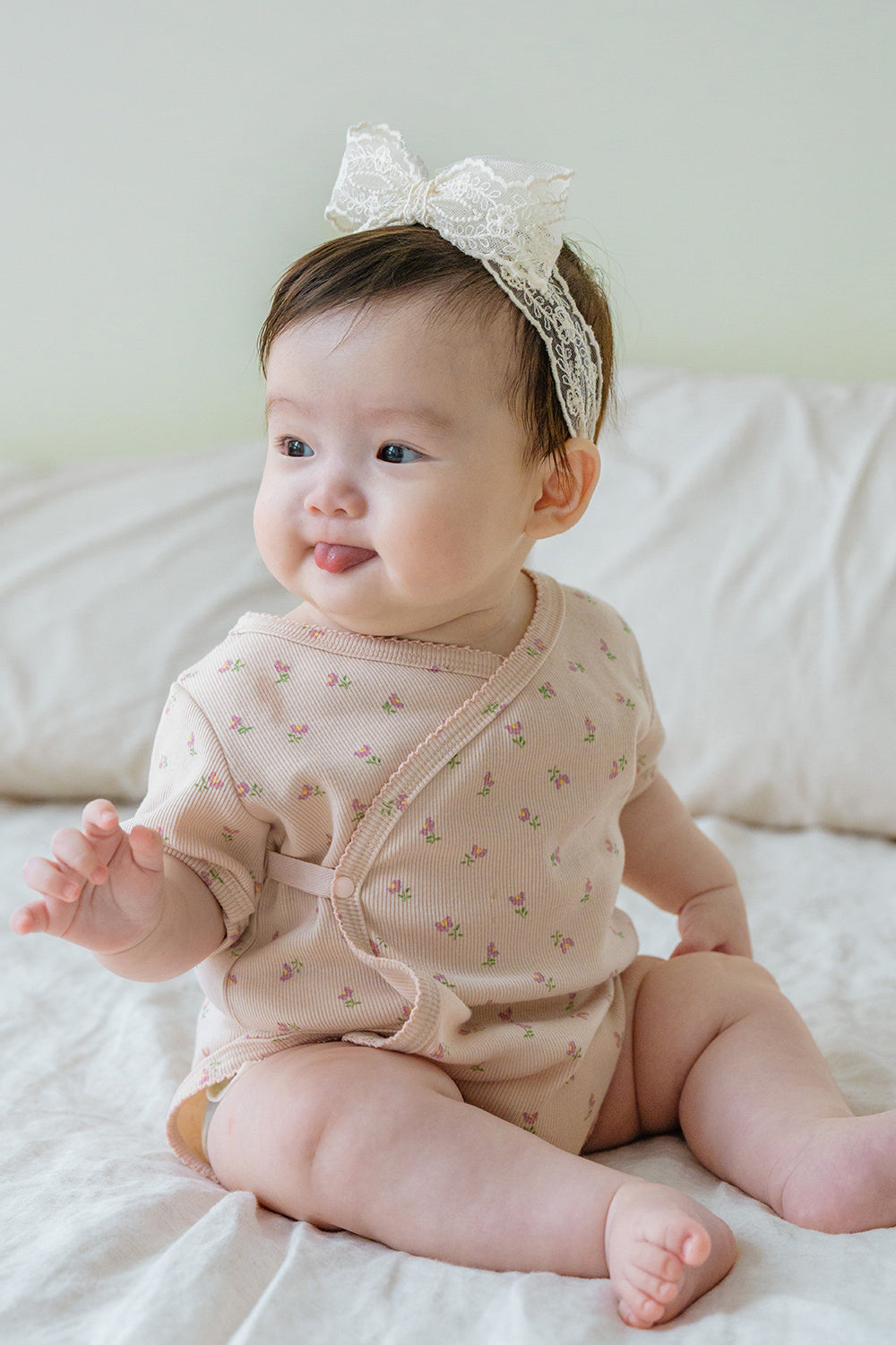 Baby girl sitting on a bed wearing Avauma Rumiere Ribbon Band and floral bodysuit, smiling with tongue out Baby girl sitting on a bed wearing Avauma Rumiere Ribbon Band and floral bodysuit, smiling with tongue out