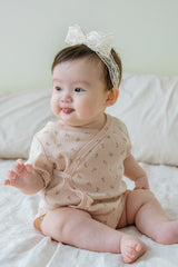 Baby girl sitting on a bed wearing Avauma Rumiere Ribbon Band and floral bodysuit, smiling with tongue out Baby girl sitting on a bed wearing Avauma Rumiere Ribbon Band and floral bodysuit, smiling with tongue out