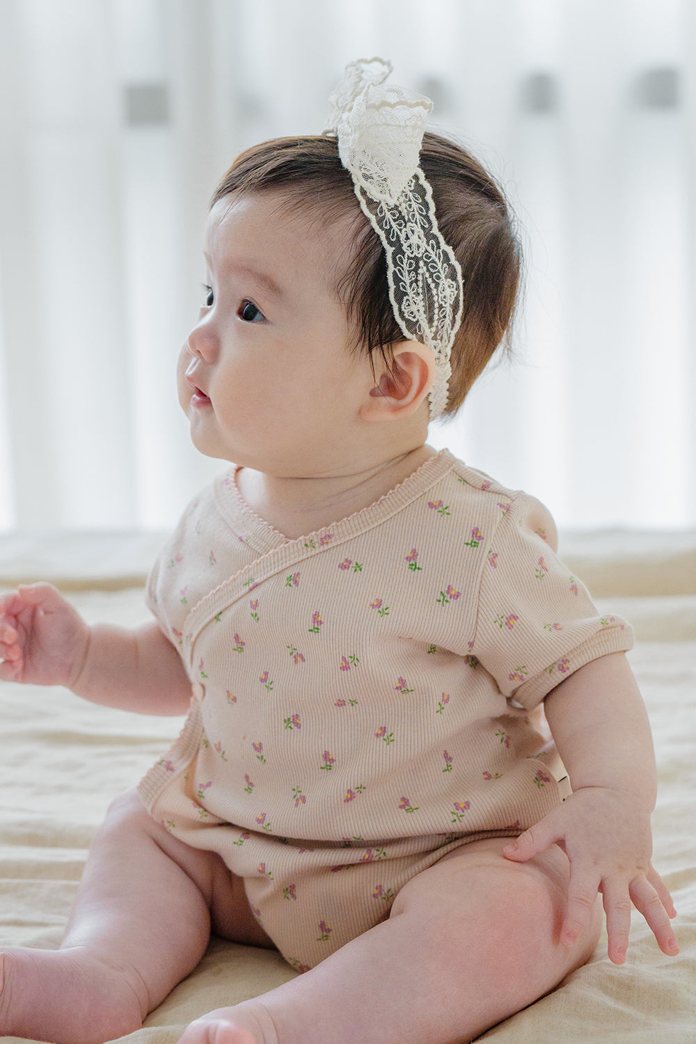 Side profile of baby girl in Avauma Rumiere Ribbon Band and peach flower-print wrap bodysuit, sitting on beige bedding Side profile of baby girl in Avauma Rumiere Ribbon Band and peach flower-print wrap bodysuit, sitting on beige bedding