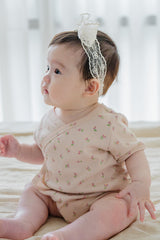 Side profile of baby girl in Avauma Rumiere Ribbon Band and peach flower-print wrap bodysuit, sitting on beige bedding Side profile of baby girl in Avauma Rumiere Ribbon Band and peach flower-print wrap bodysuit, sitting on beige bedding