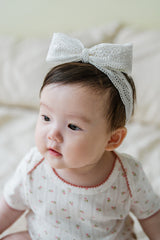 Baby girl wearing Avauma Rumiere Ribbon Band in Cotton Cream with white floral baby bodysuit, sitting on a bed Baby girl wearing Avauma Rumiere Ribbon Band in Cotton Cream with white floral baby bodysuit, sitting on a bed