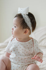 Side view of baby girl in Avauma Rumiere Ribbon Band - Cotton Cream and soft floral romper, sitting on beige bedding Side view of baby girl in Avauma Rumiere Ribbon Band - Cotton Cream and soft floral romper, sitting on beige bedding