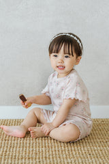 A young girl sitting on the floor, smiling, wearing Avauma Sienna Rose short sleeve set in Rosy Powder with pink floral prints. A young girl sitting on the floor, smiling, wearing Avauma Sienna Rose short sleeve set in Rosy Powder with pink floral prints.