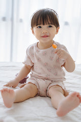 A baby girl sitting on a bed, wearing Avauma Sienna Rose short sleeve set in Rosy Powder, with a pink floral pattern and wavy hems. A baby girl sitting on a bed, wearing Avauma Sienna Rose short sleeve set in Rosy Powder, with a pink floral pattern and wavy hems.