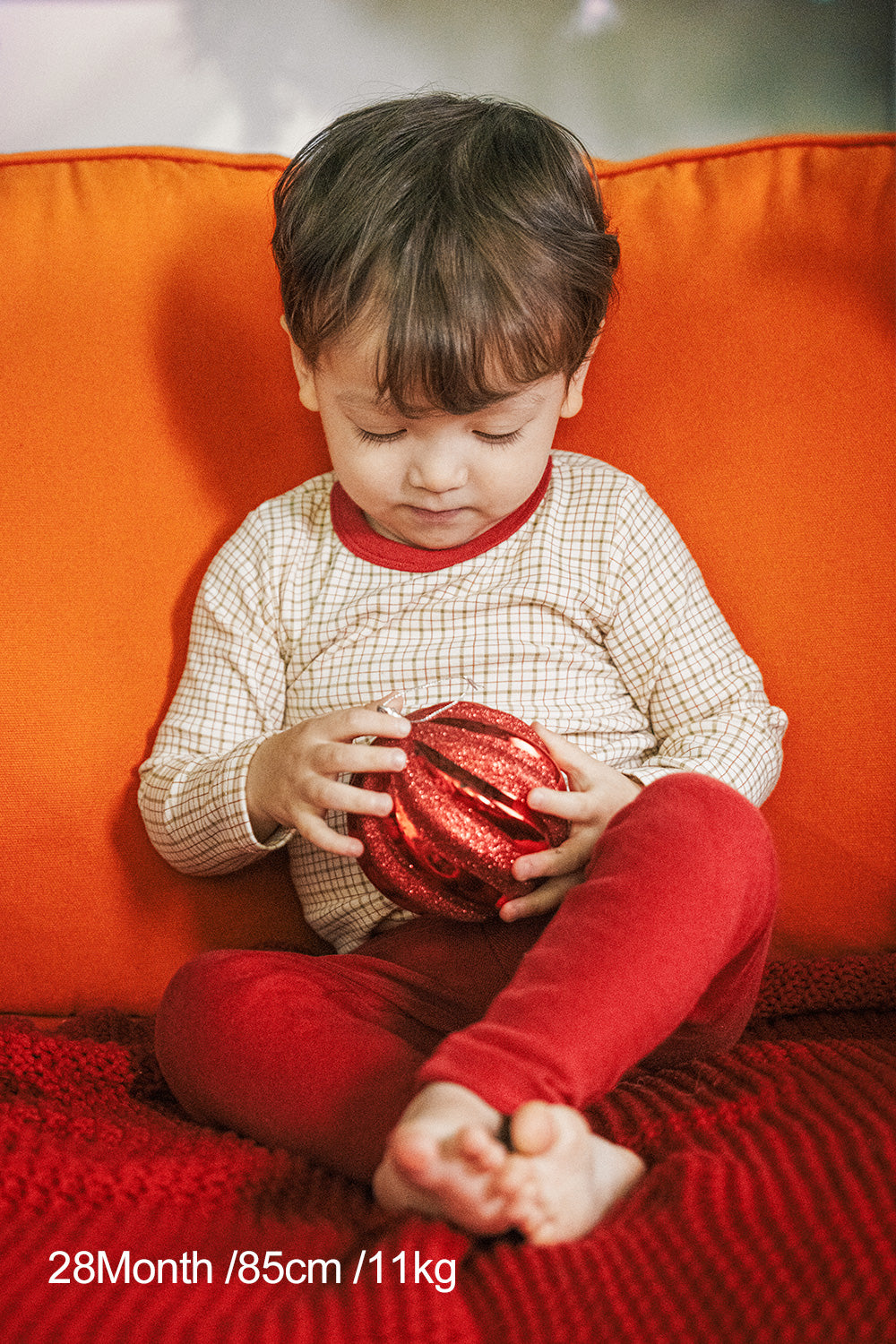 Toddler in Avauma Holiday Long Sleeve Pajama Set - Cranberry Check, holding a red Christmas ornament on a cozy orange couch