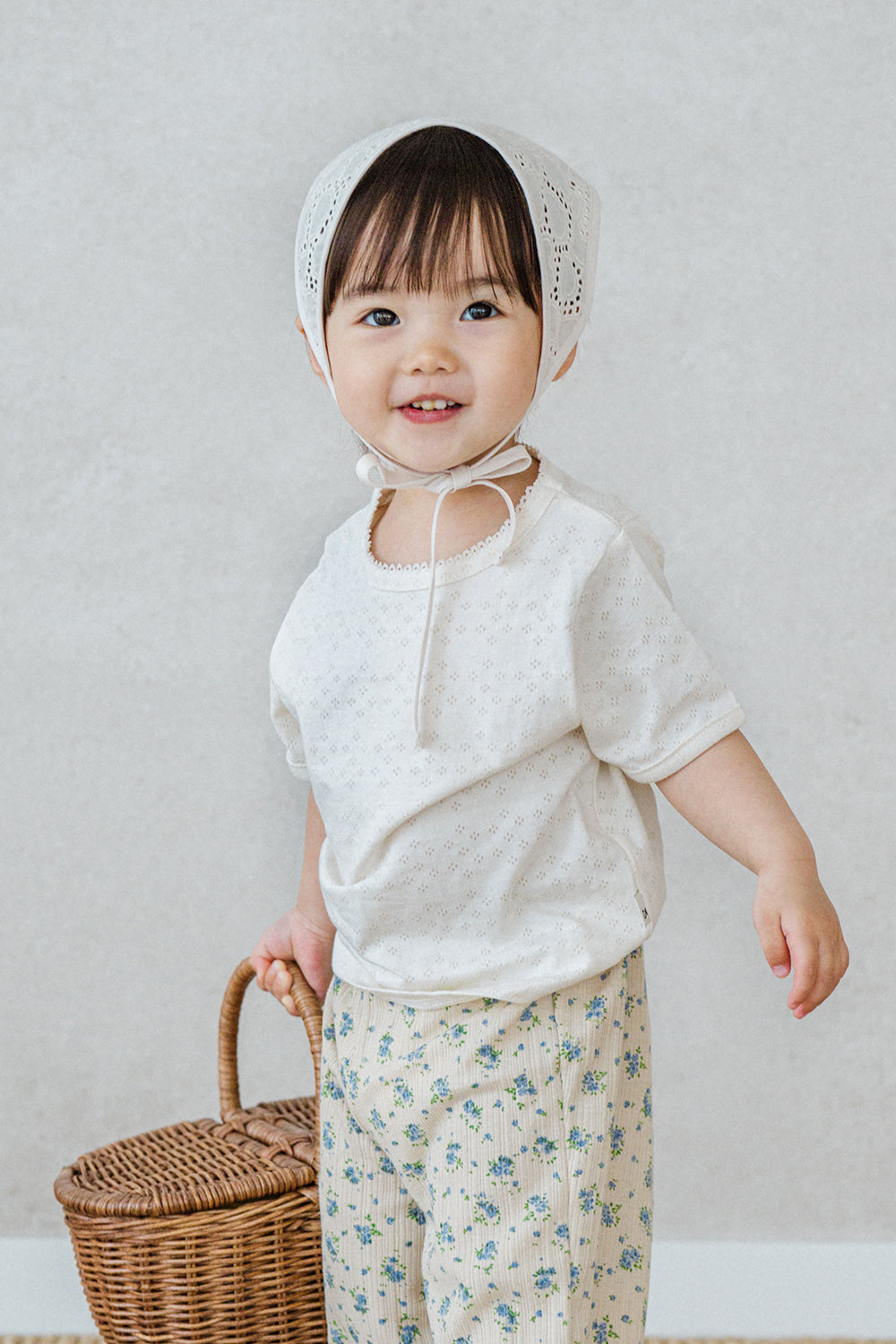 Toddler girl wearing Avauma milk pearl lace short sleeve top and floral pants, holding a wicker basket

