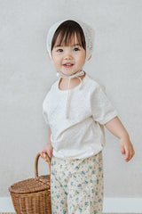 Toddler girl wearing Avauma milk pearl lace short sleeve top and floral pants, holding a wicker basket

