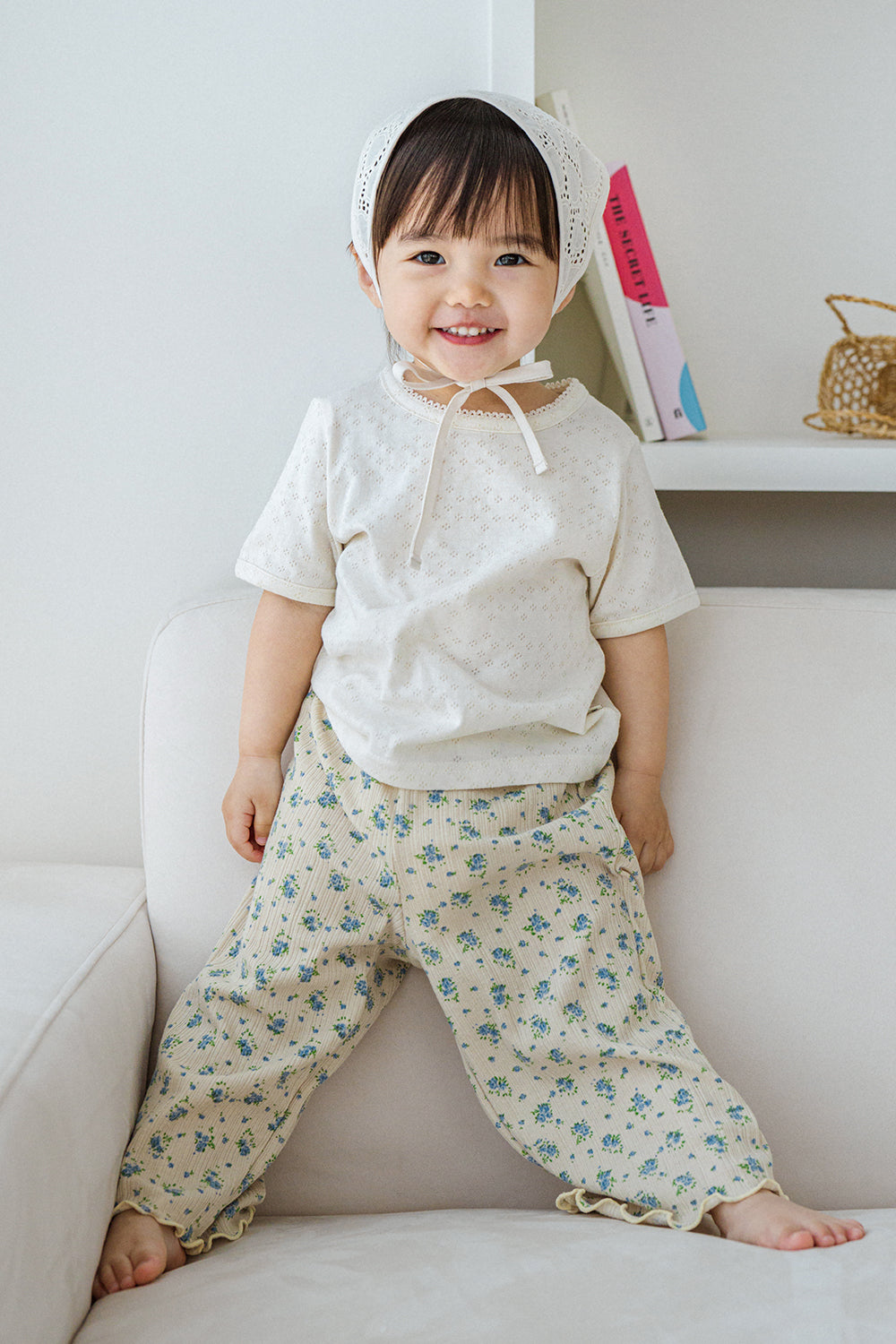 Smiling toddler in Avauma milk pearl lace short sleeve tee and flower print pants, standing on sofa

