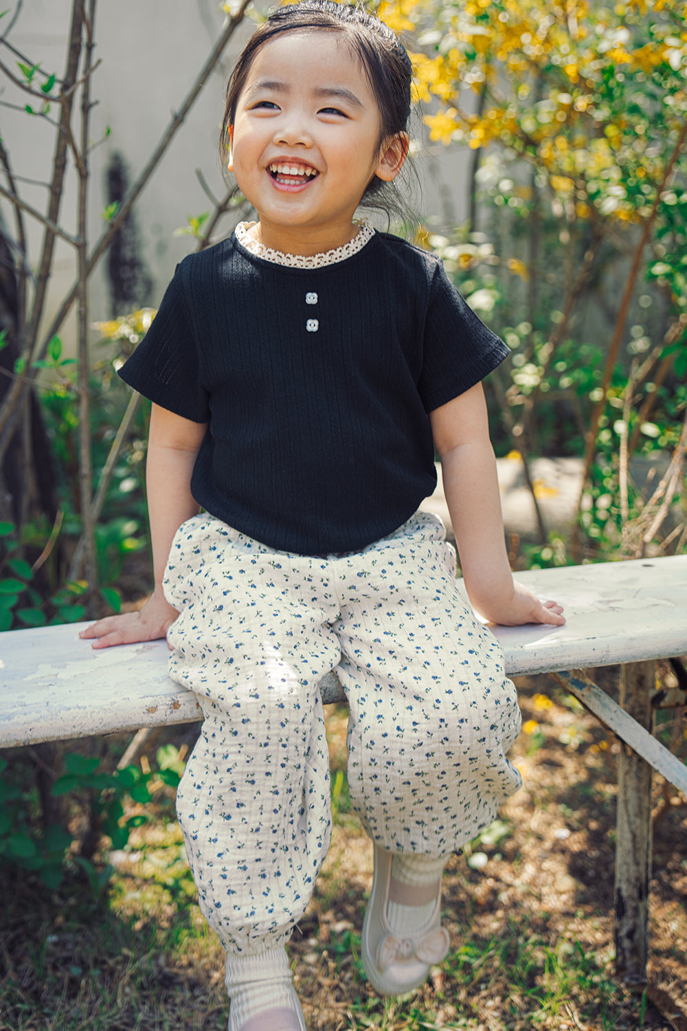 Happy toddler girl sitting on a bench wearing stylish Avauma 100% cotton muslin pants and black ribbed shirt, casual kids fashion.