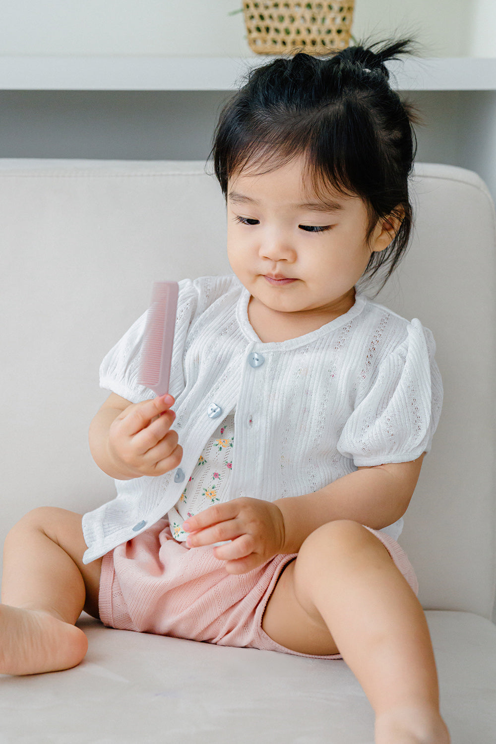 Toddler sitting on sofa wearing Avauma Buttercumin cotton ivory cardigan and pink shorts.

