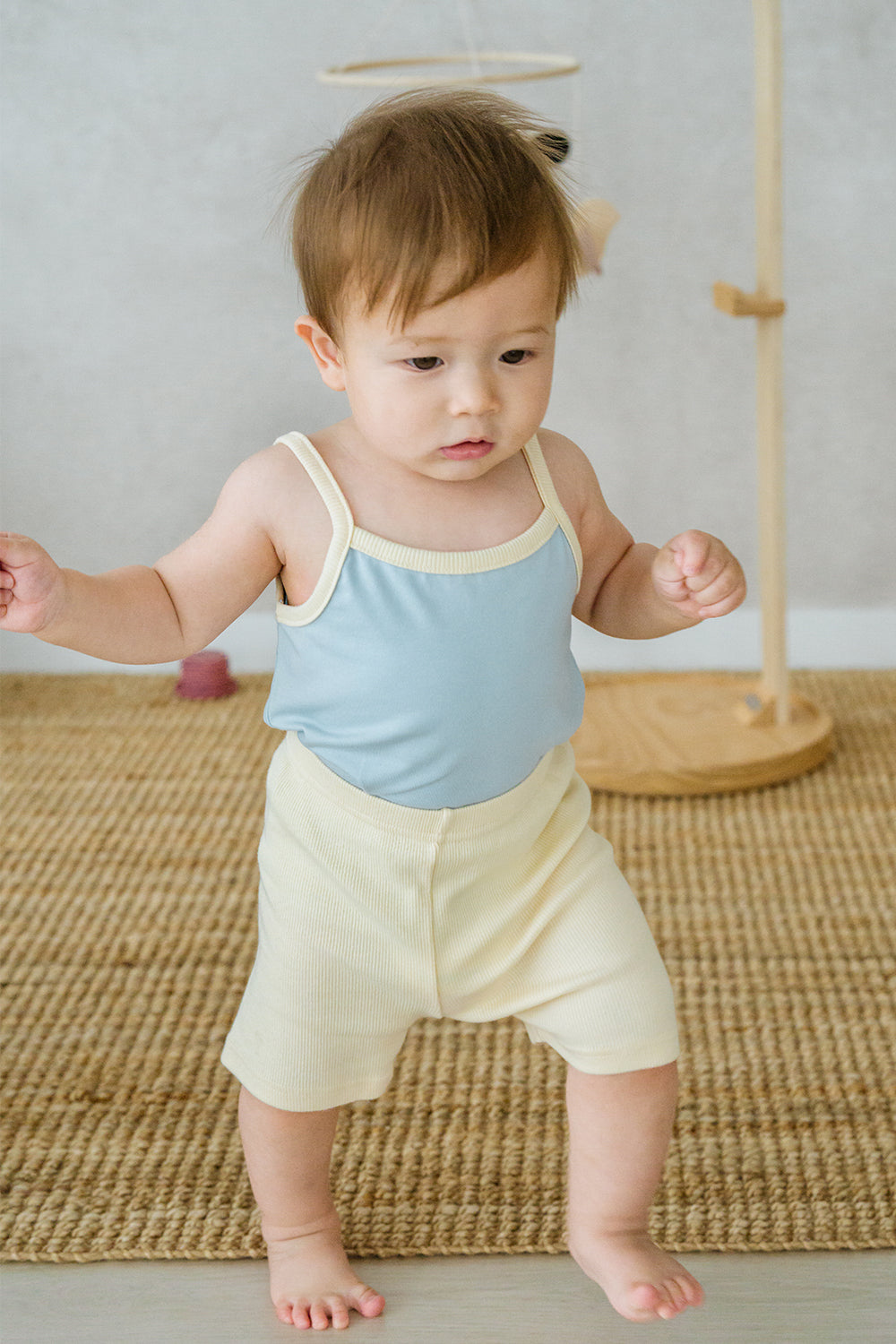 Baby wearing Avauma pale lime sleeveless set, standing on woven mat
