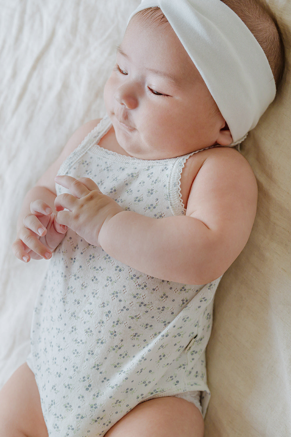 Sweet baby in Avauma Lily Sleeveless Bodysuit Cream Blue. Newborn lying down with closed eyes, wearing a cream and blue floral print romper and white headband.

