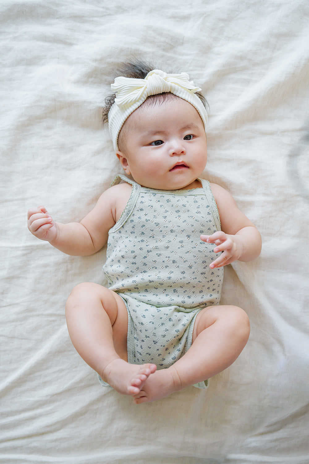 Adorable baby model in Avauma Lily Sleeveless Bodysuit Sage Flower. Infant lying down in a soft sage green floral print bodysuit with a twisted white headband.