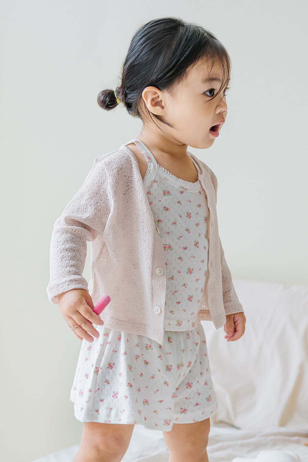 Toddler girl wearing avauma Little Shell Cardigan – Almond Cream over a floral dress, standing indoors.