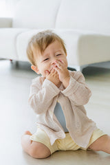 Baby sitting and smiling while wearing Avauma Littleshell cardigan in almond cream over a beige romper.

