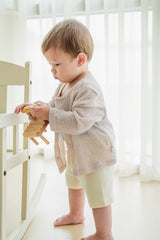 Baby boy standing with plush toy, wearing Avauma Littleshell cardigan in almond cream.

