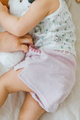 Infant lying on their side in Avauma Pocket Day Shorts Milk Pink. Baby wearing soft pink shorts and a white top, cuddling a white plush toy on a bed.
