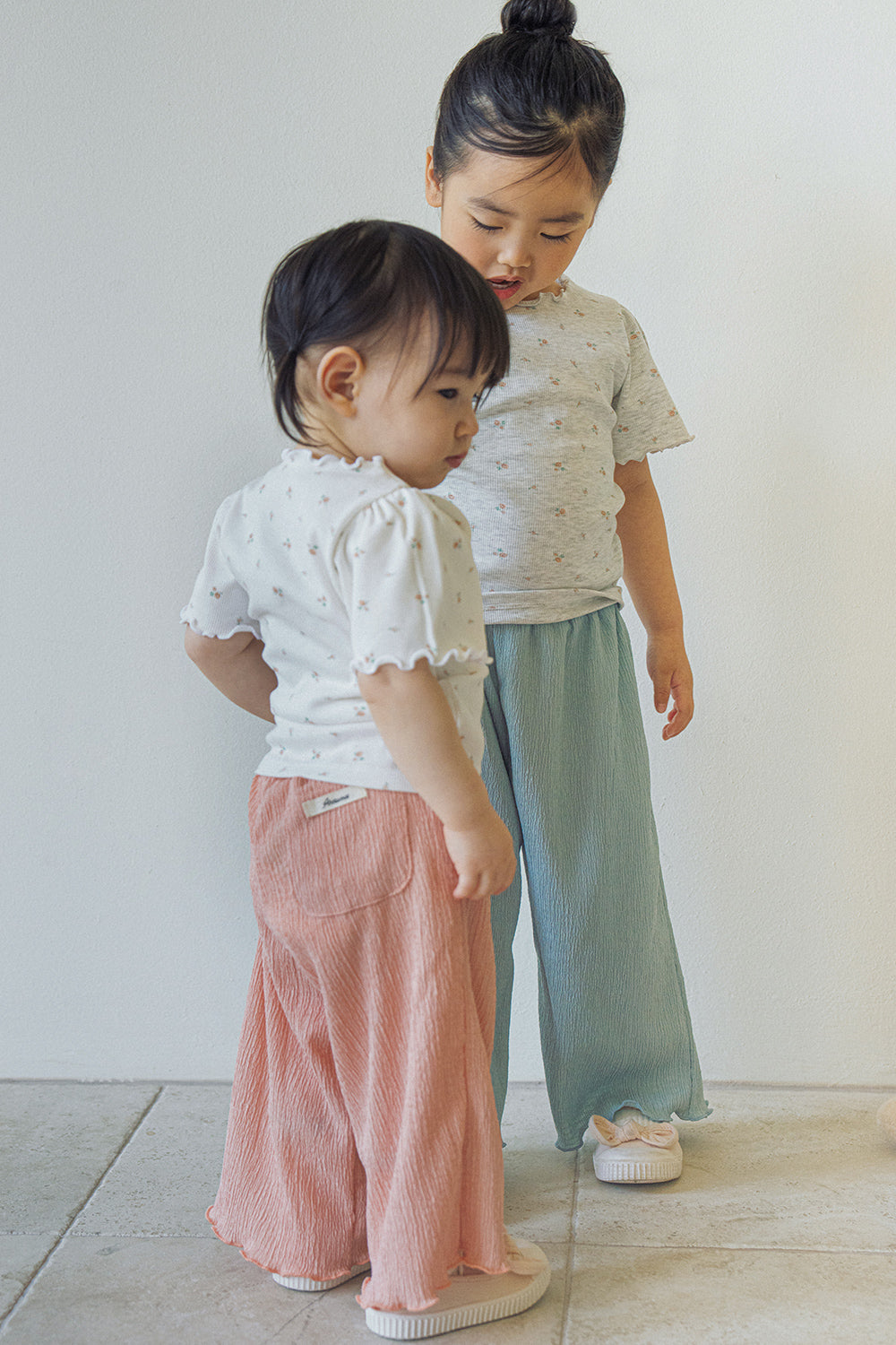 Two young girls modeling Avauma summer outfits; one in the cream floral top and pink pants, the other in a similar grey top and light blue pants.