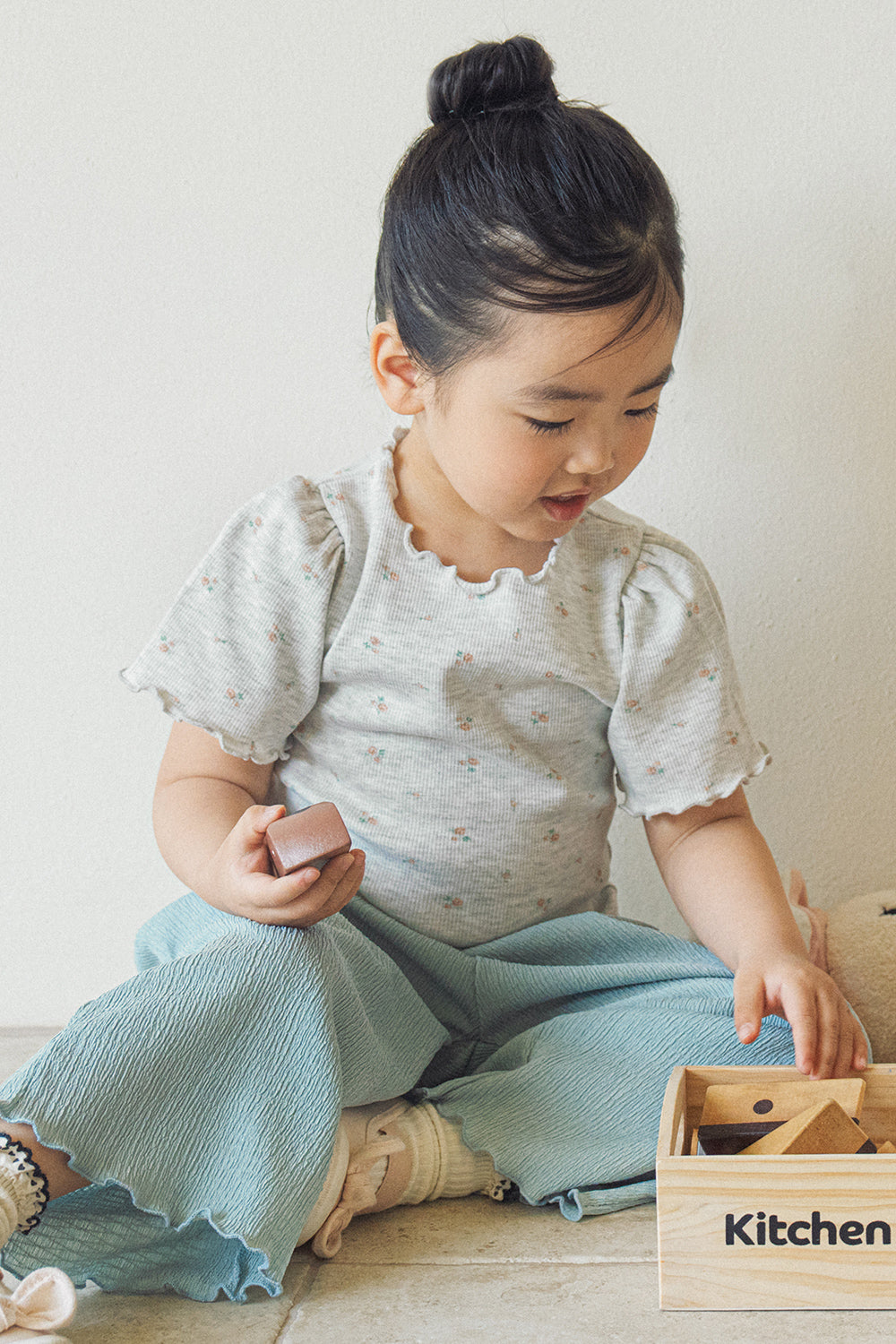 A young girl sitting and playing while wearing the Avauma grey floral tee paired with teal wide-leg ribbed pants.