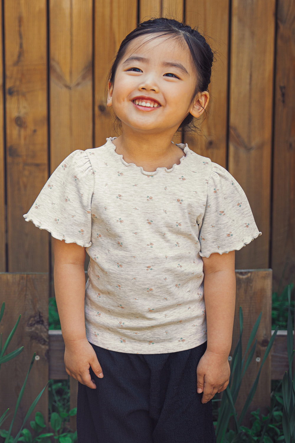Smiling toddler modeling the Avauma summer floral t-shirt and black relaxed-fit trousers in an outdoor garden setting.