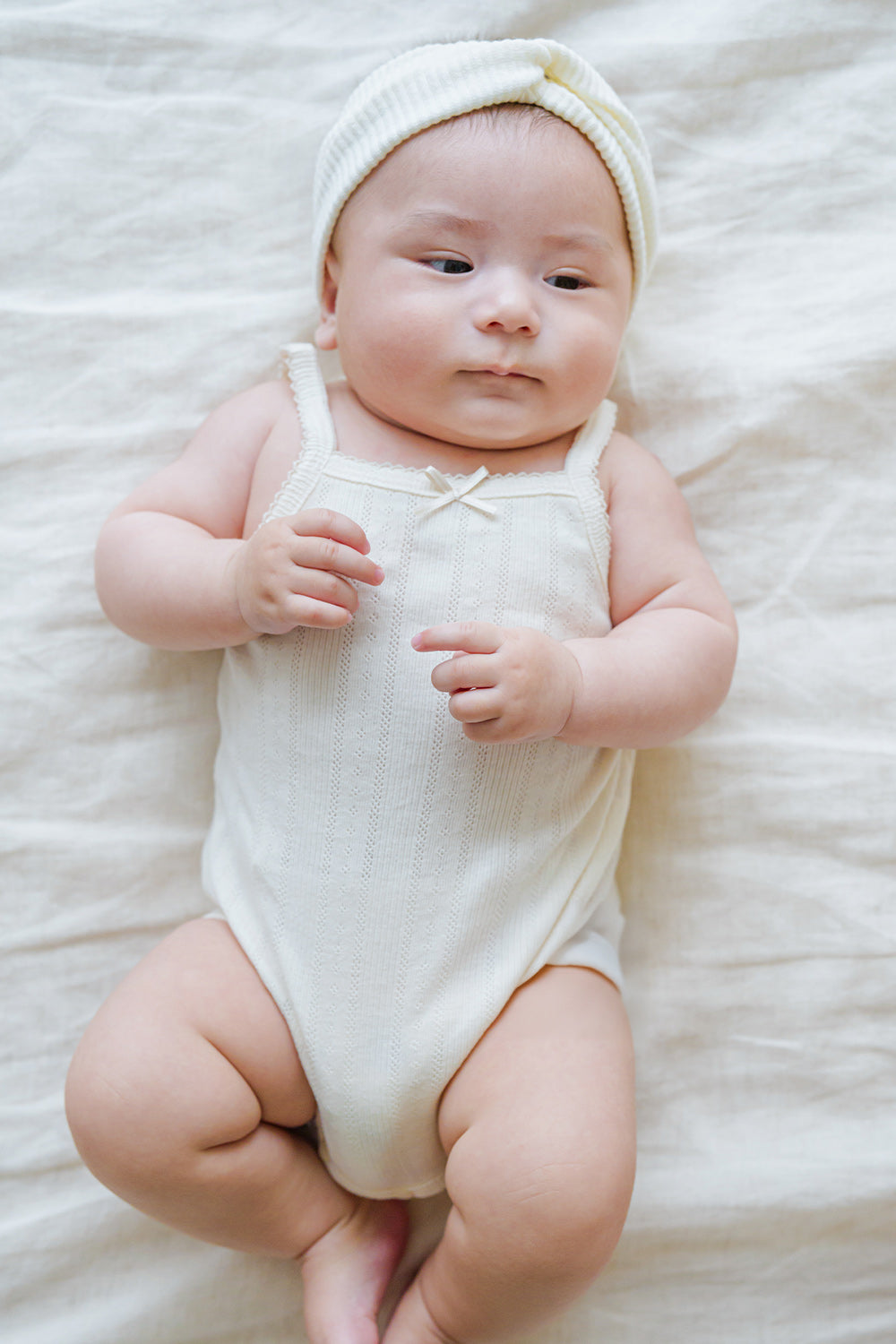 Cute newborn in Avauma Summer Picot Sleeveless Bodysuit Buttermilk. Baby lying down in a soft white ribbed romper with a delicate bow detail and matching headband.