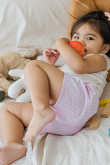 Toddler lying down in Avauma Sunny Stripe Pants Lotus Pink. Child holding a fruit, wearing light pink ribbed shorts and a white top.

