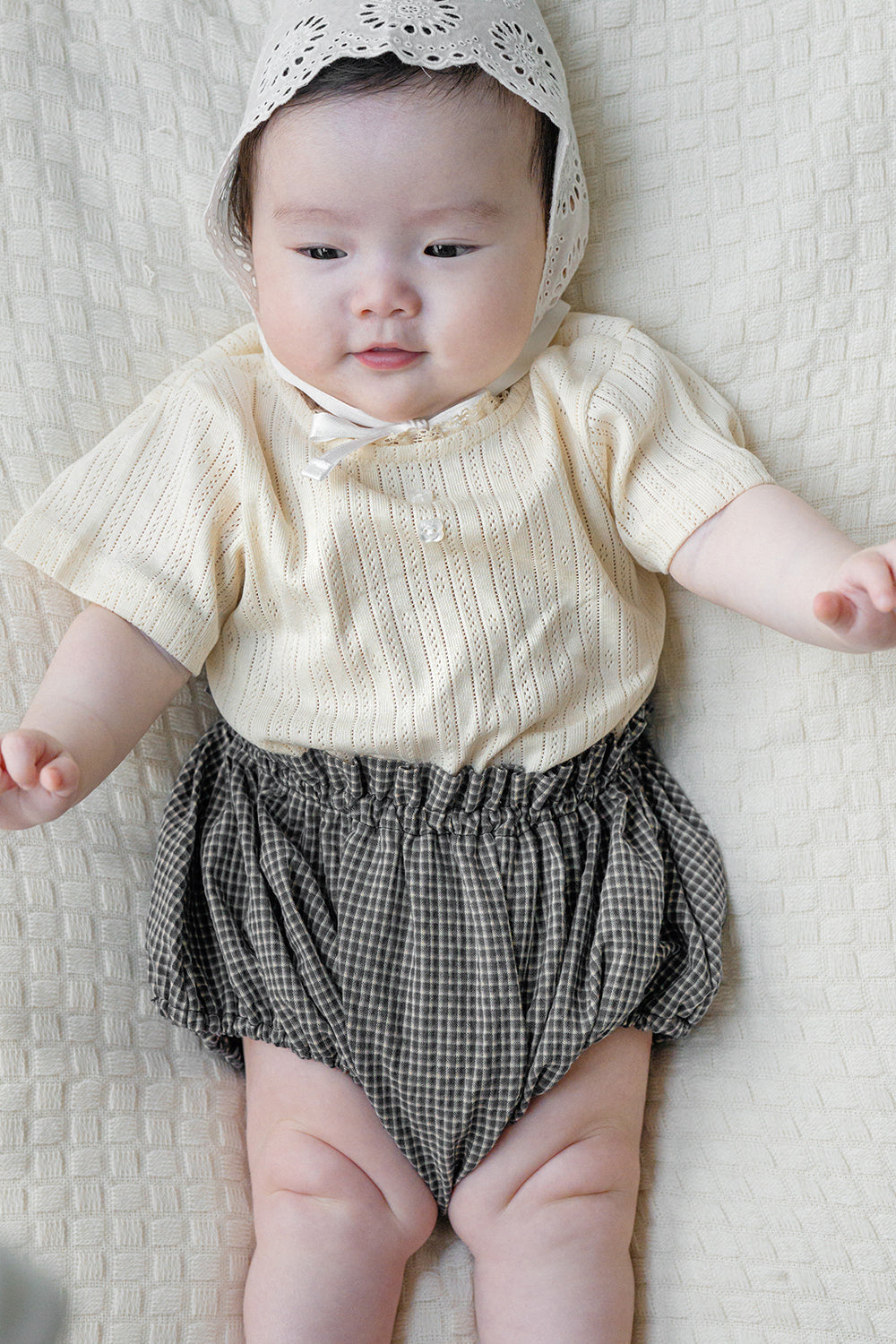 Baby wearing Avauma Berry Puff Bloomer in Camellia Check, lying on textured cream bedding